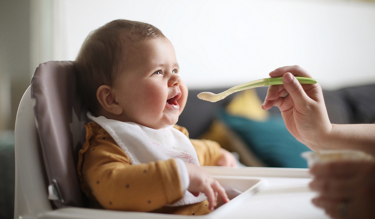 A baby in a highchair opens it's mouth to be fed from a baby spoon.