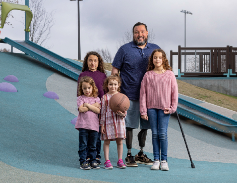 TIRR Memorial Hermann Osseointegration patient, Alex, stands with his four daughters at a play ground.