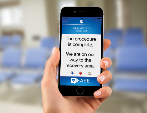 A hand holds up a mobile phone with a message about a loved one's procedure; the background is a hospital waiting room.