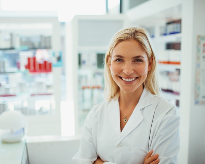 A female pharmacist smiles in front of the pharmacy stock.
